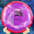 Load image into Gallery viewer, Top-down view of a purple Axiom Time Lapse distance driver disc golf disc with a red swirl rim in Neutron plastic featuring a silver holo and black foil Simon Line stamp.
