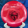 Load image into Gallery viewer, Top-down view of a brand-new metallic red Mint Discs Alpha fairway driver disc golf disc in Sublime plastic with a red foil Unearthed serial run SB-AL07-24 graphic stamp.
